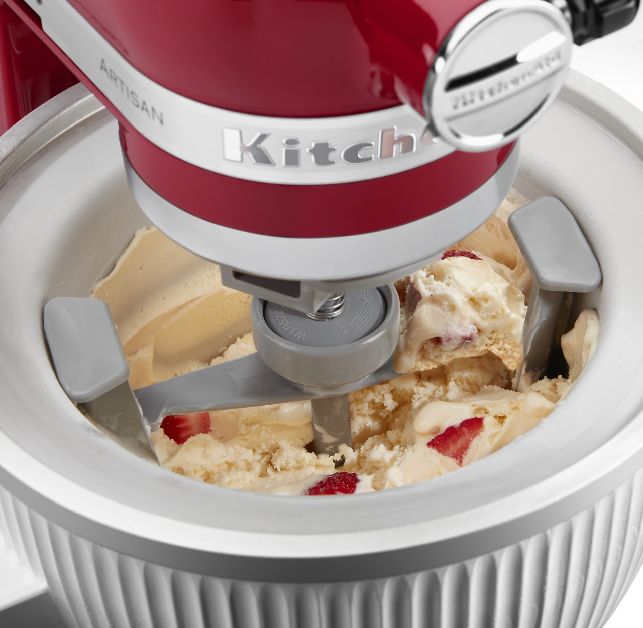 Eine rote KitchenAid Artisan Eismaschine rührt Vanilleeis mit Erdbeerstücken. Die Maschine steht auf einer weißen Oberfläche.