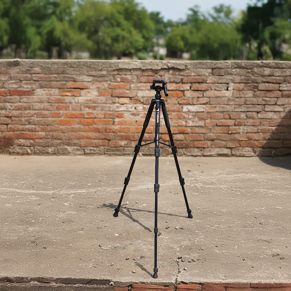 Siyah bir tripod, tuğla bir duvarın önünde beton bir yüzeyde duruyor. Arka planda yeşil ağaçlar var.