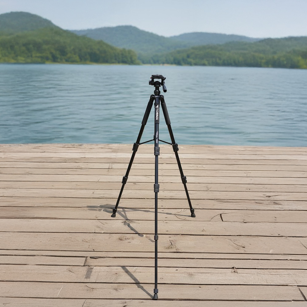 Siyah bir tripod, ahşap bir iskelede duruyor, arka planda sakin bir göl ve uzaktaki yeşil tepeler var.