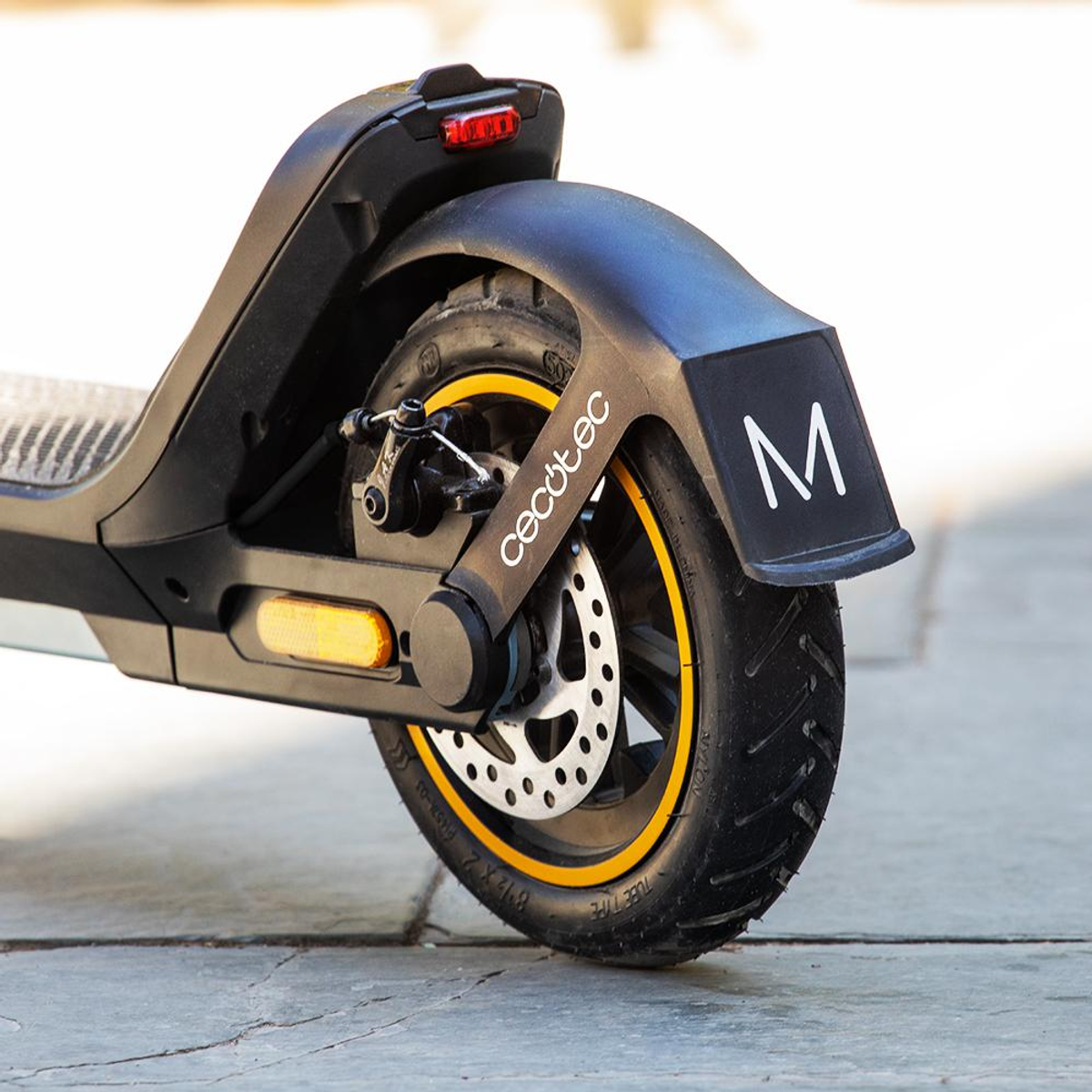 Czarna tylna opona hulajnogi elektrycznej z żółtymi akcentami. Opona jest czarna. Błotnik ma białą literę 'M'. Tło jest szare.
