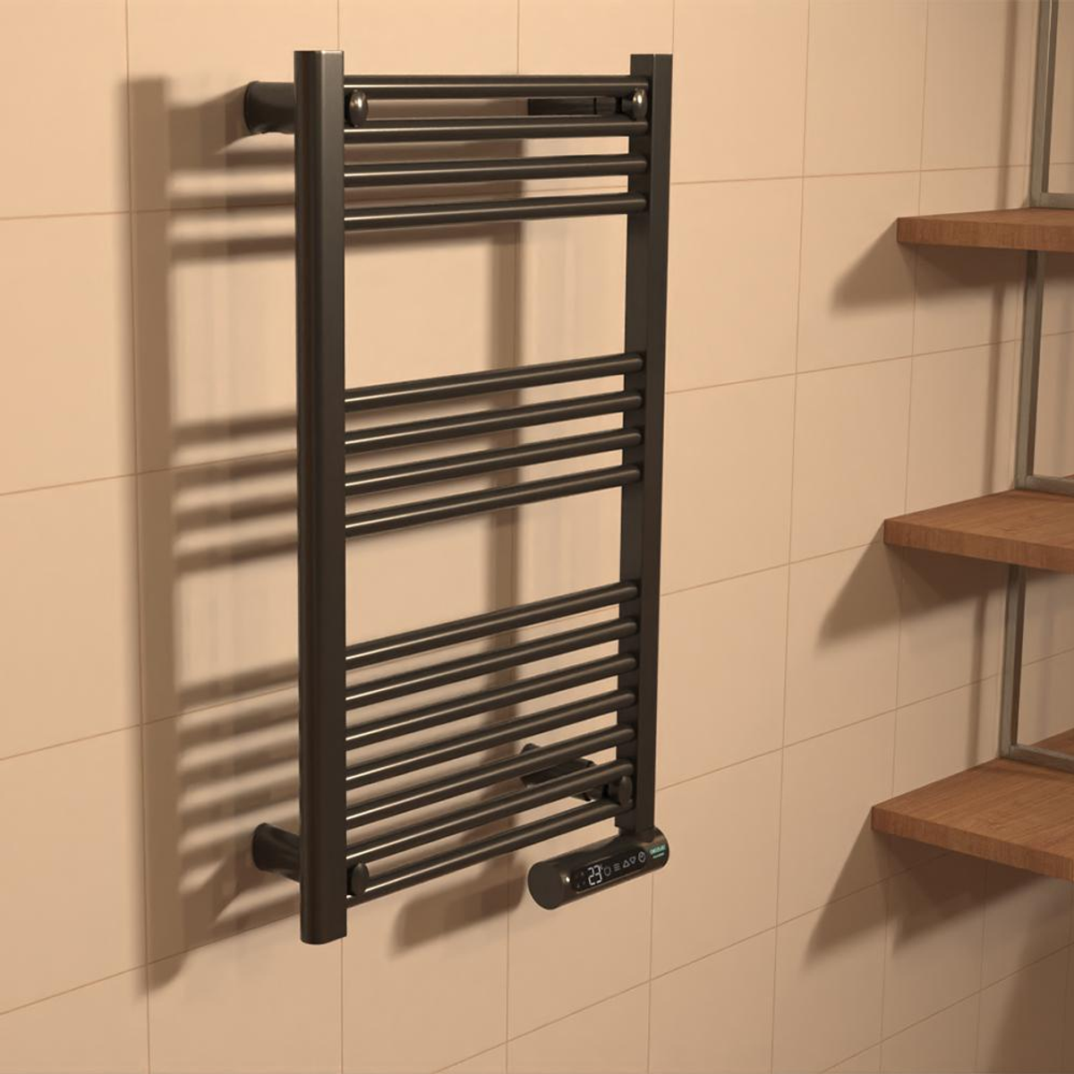 Ciemnoszary podgrzewacz do ręczników zamontowany na beżowej, kafelkowanej ścianie. Dwie drewniane półki po prawej stronie. Podgrzewacz posiada wyświetlacz cyfrowy.