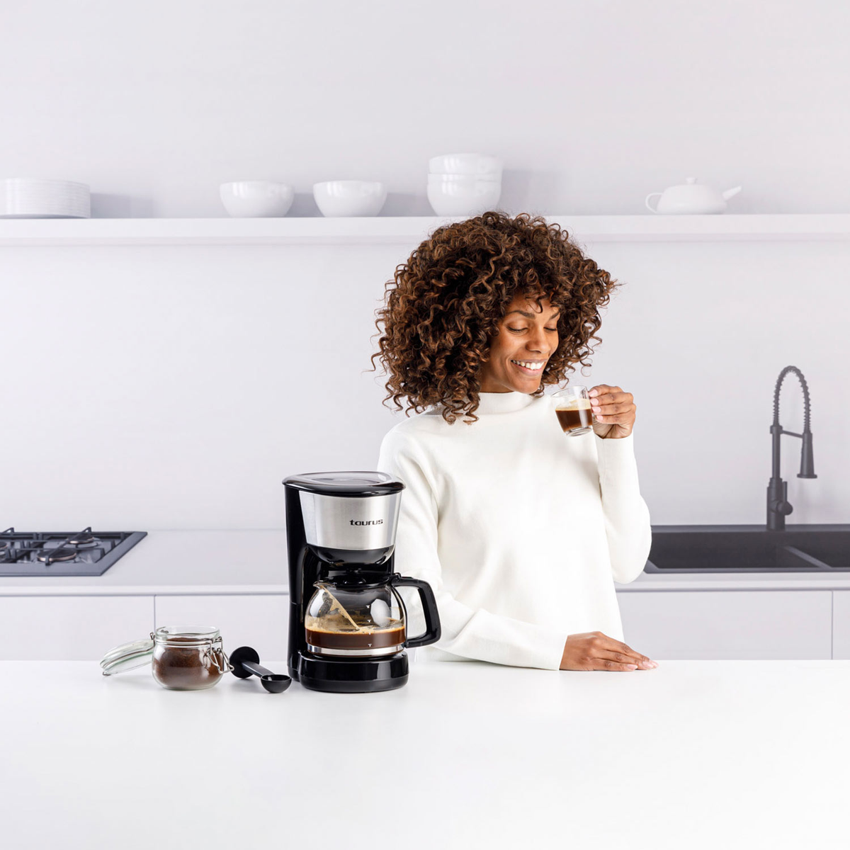 Kobieta w białym swetrze uśmiecha się, trzymając kawę. Ekspres do kawy, słoik i łyżka na blacie. Tło kuchni.