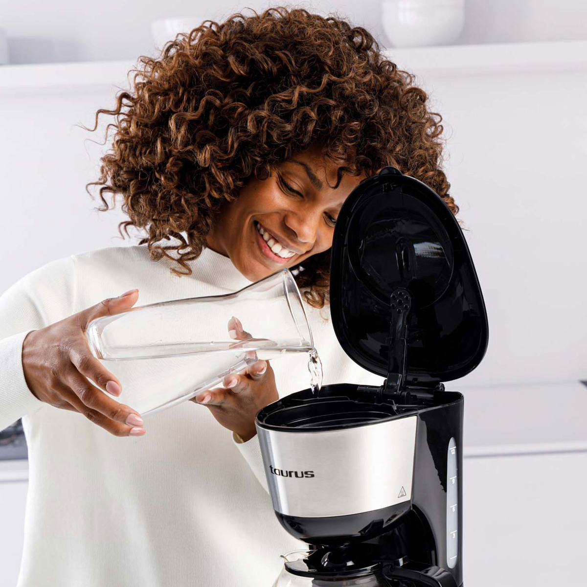 Kobieta wlewa wodę ze szklanej karafki do czarno-srebrnego ekspresu do kawy. Uśmiecha się, z kręconymi brązowymi włosami.