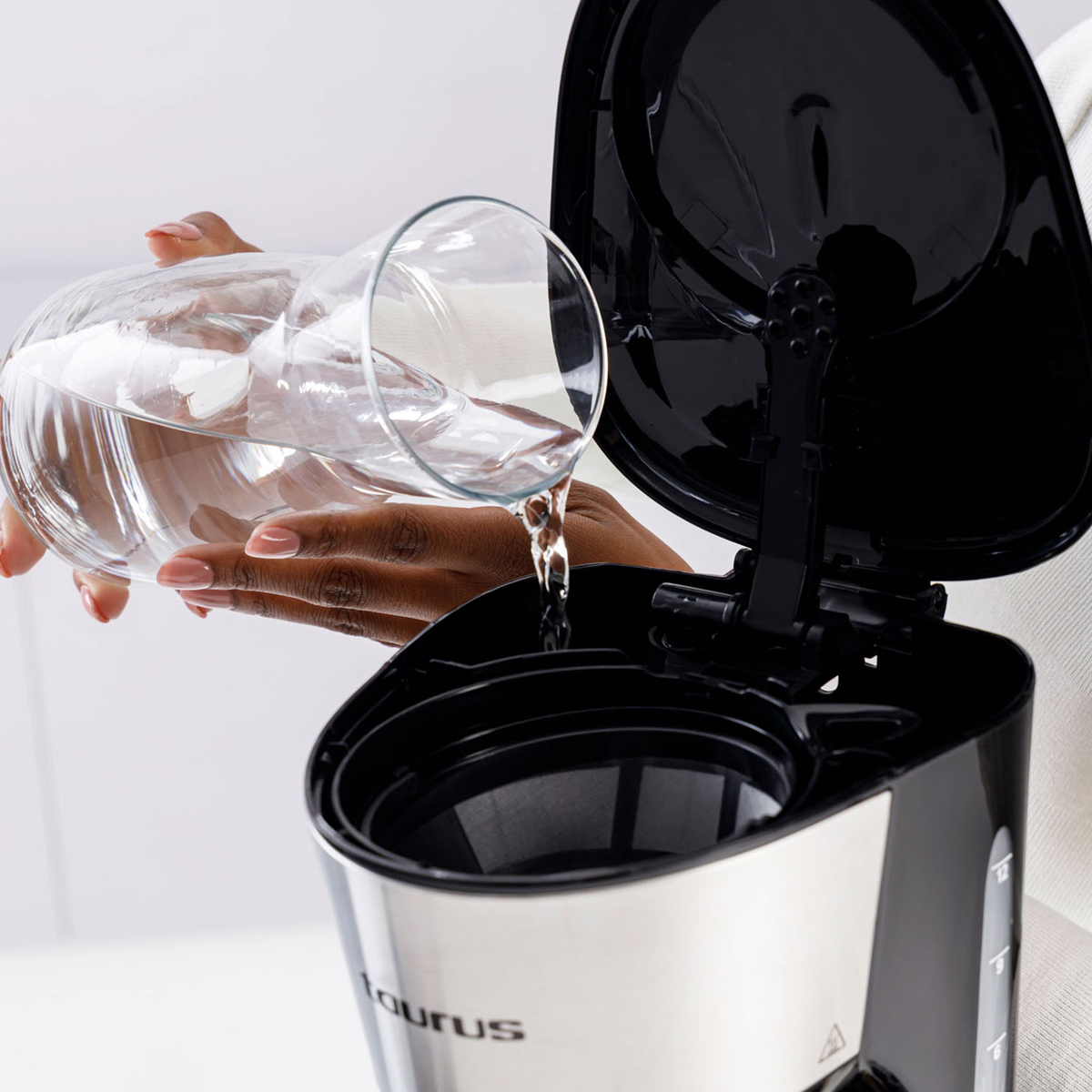 Osoba wlewa wodę ze szklanej karafki do czarno-srebrnego ekspresu do kawy. Pokrywa ekspresu do kawy jest otwarta.
