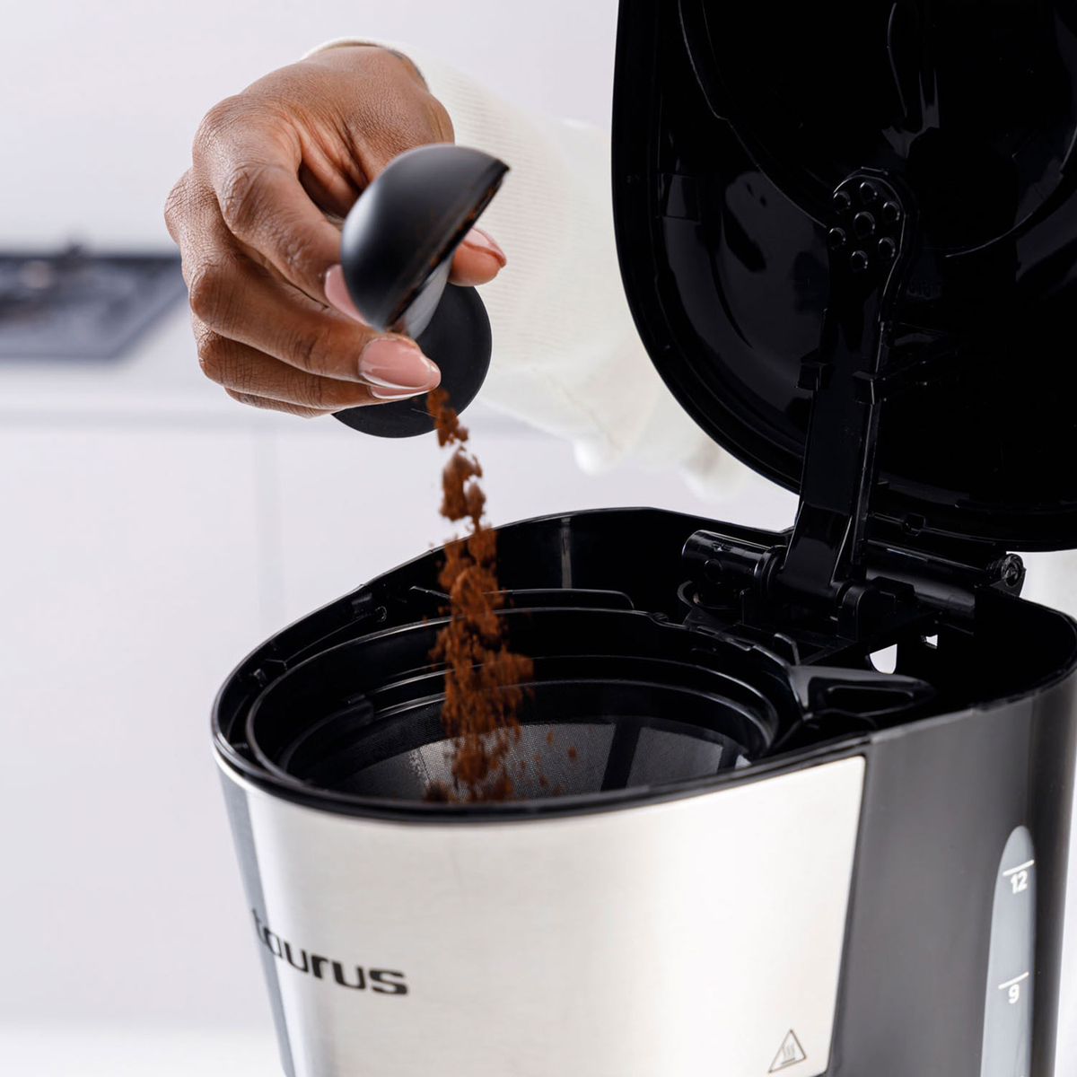 Ręka wsypuje zmieloną kawę z miarki do czarno-srebrnego ekspresu do kawy. Pokrywa maszyny jest otwarta.