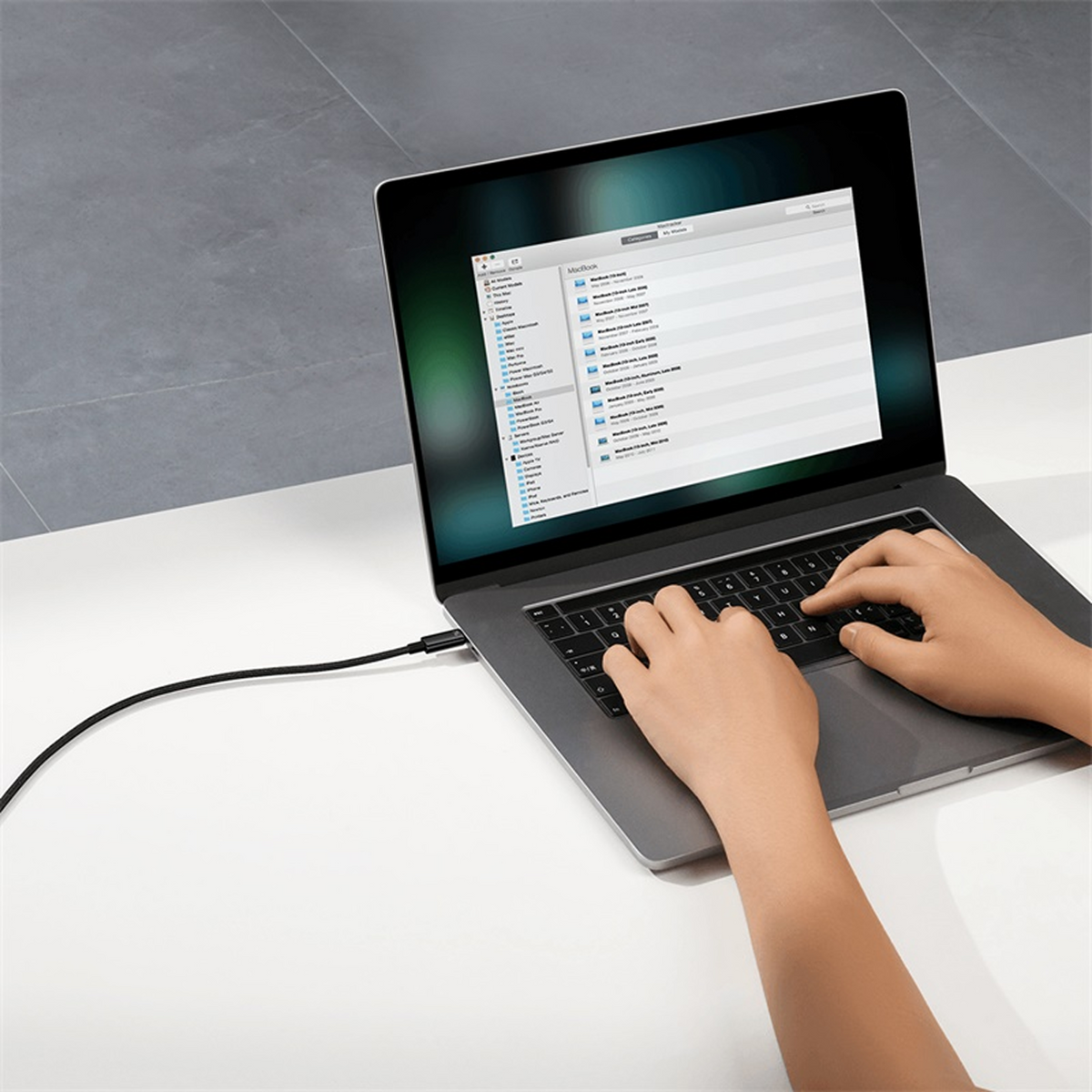 Laptop z podłączonym czarnym kablem. Ręce piszą na klawiaturze. Ekran wyświetla katalog plików.
