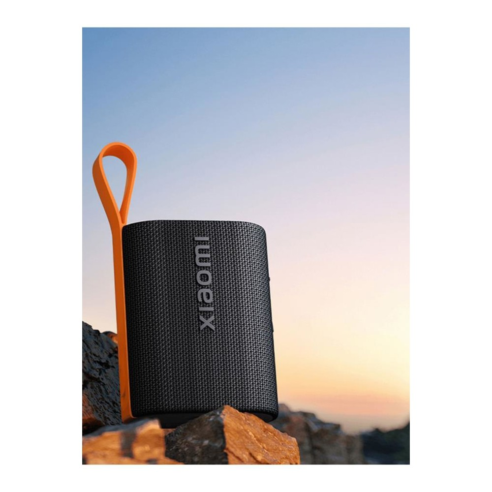 Czarny głośnik Xiaomi z pomarańczowym uchwytem w kształcie pętli stoi na skałach na tle zachodzącego słońca. Nazwa głośnika jest widoczna.