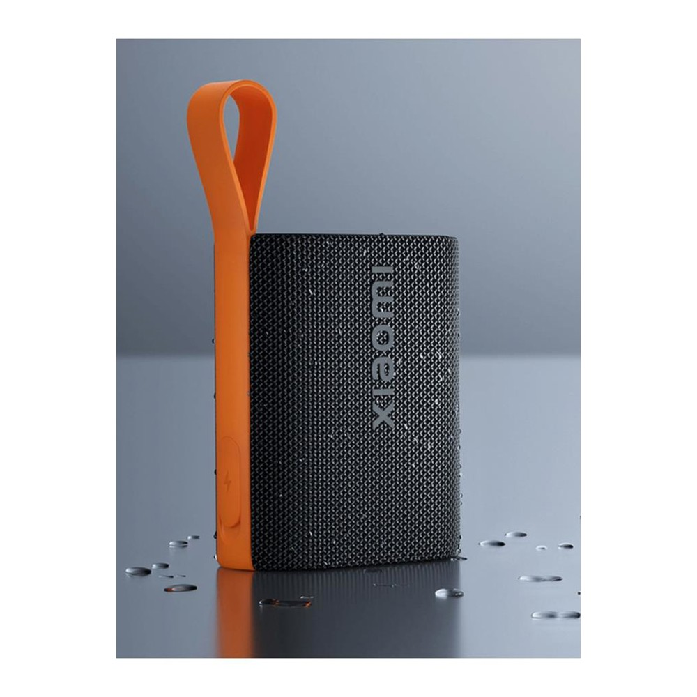 Czarny głośnik Xiaomi z pomarańczowym uchwytem. Krople wody na powierzchni. Szare tło. Głośnik jest prostokątny.