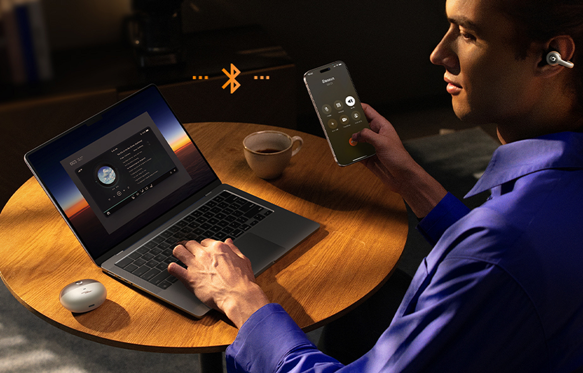 Ein Mann benutzt Laptop und Smartphone, trägt Ohrhörer. Kaffeetasse und Ladecase auf dem Tisch. Bluetooth-Symbol.