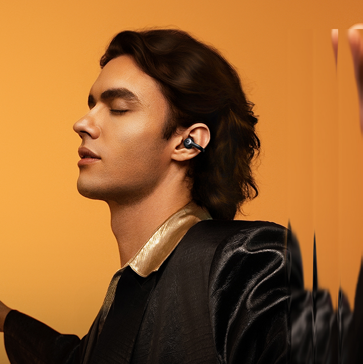 Ein Mann mit langen Haaren, der einen Ohrhörer trägt, die Augen geschlossen, in einer schwarzen Jacke mit goldenem Besatz, orangefarbener Hintergrund.