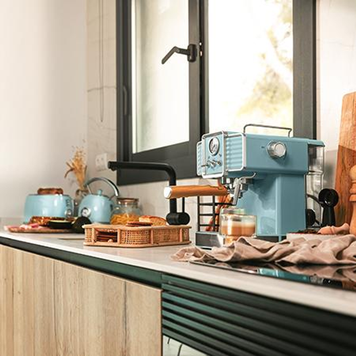 Una escena de cocina con una máquina de espresso azul, una tostadora y una tetera. Hay una ventana en el fondo con luz natural.