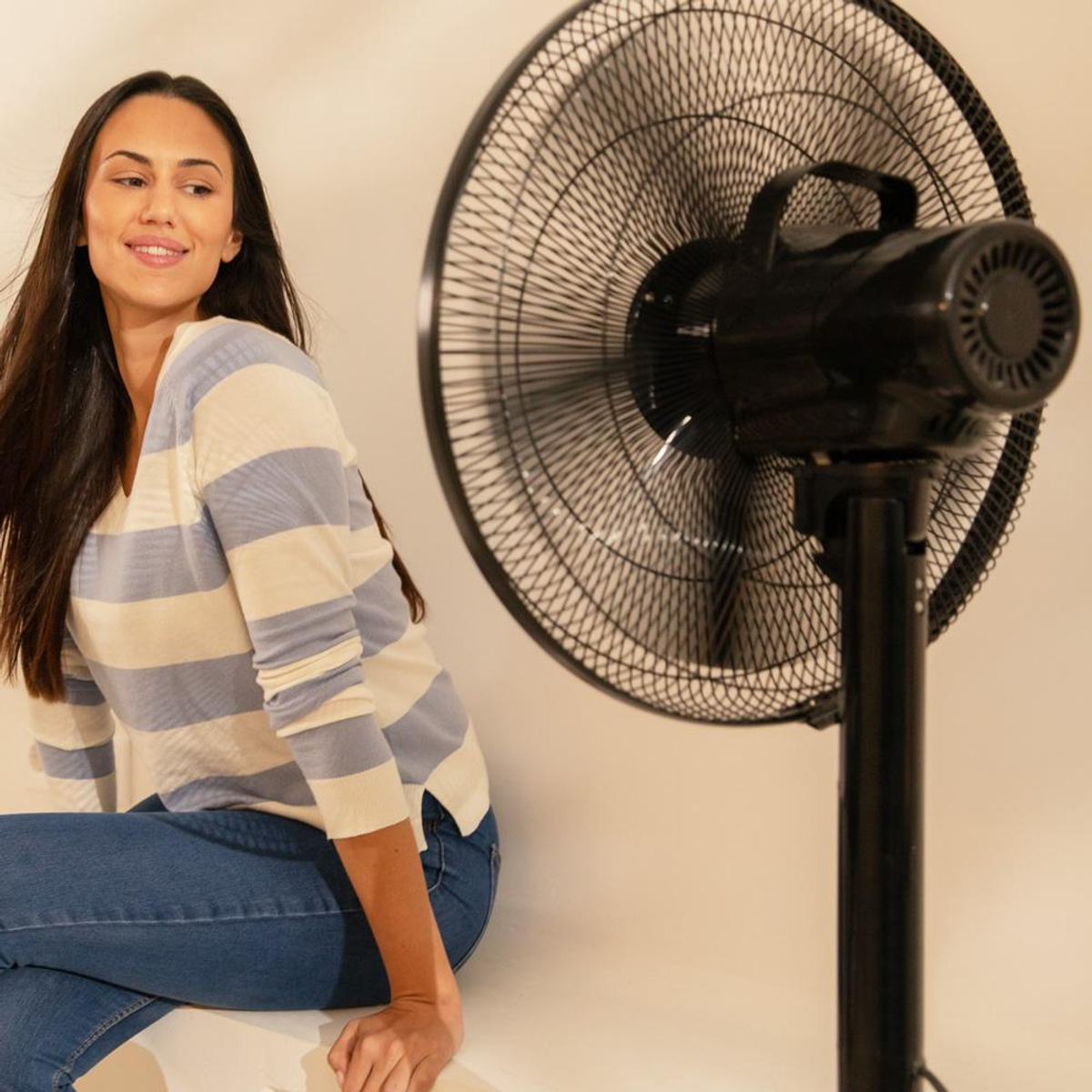 Kobieta siedzi obok czarnego wentylatora. Ma na sobie koszulę w paski i dżinsy. Widoczne są łopatki wentylatora.