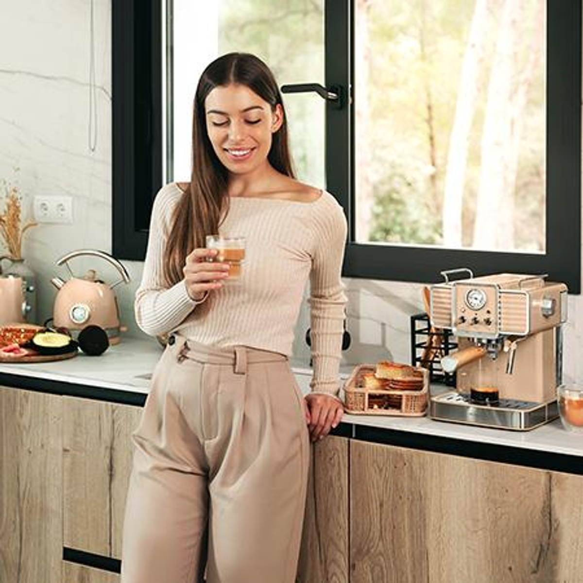 Una donna sorride, tenendo in mano una bevanda, in una cucina. C'è una macchina per il caffè, pane e un bollitore. Sono visibili mobili in legno e una finestra.