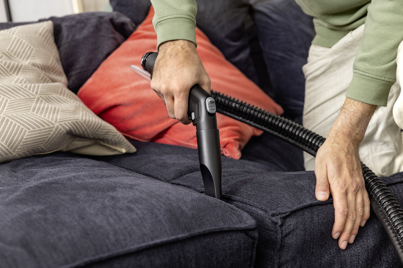 Osoba odkurza granatową sofę. W tle pomarańczowe i wzorzyste poduszki. Odkurzacz jest czarny.