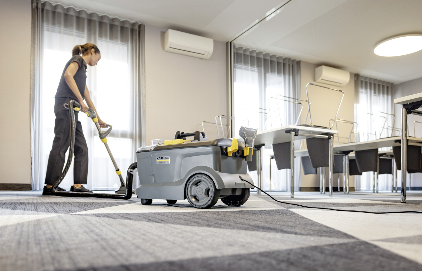 Kobieta używa odkurzacza Karcher na dywanie w pokoju. Stoły i krzesła w tle.