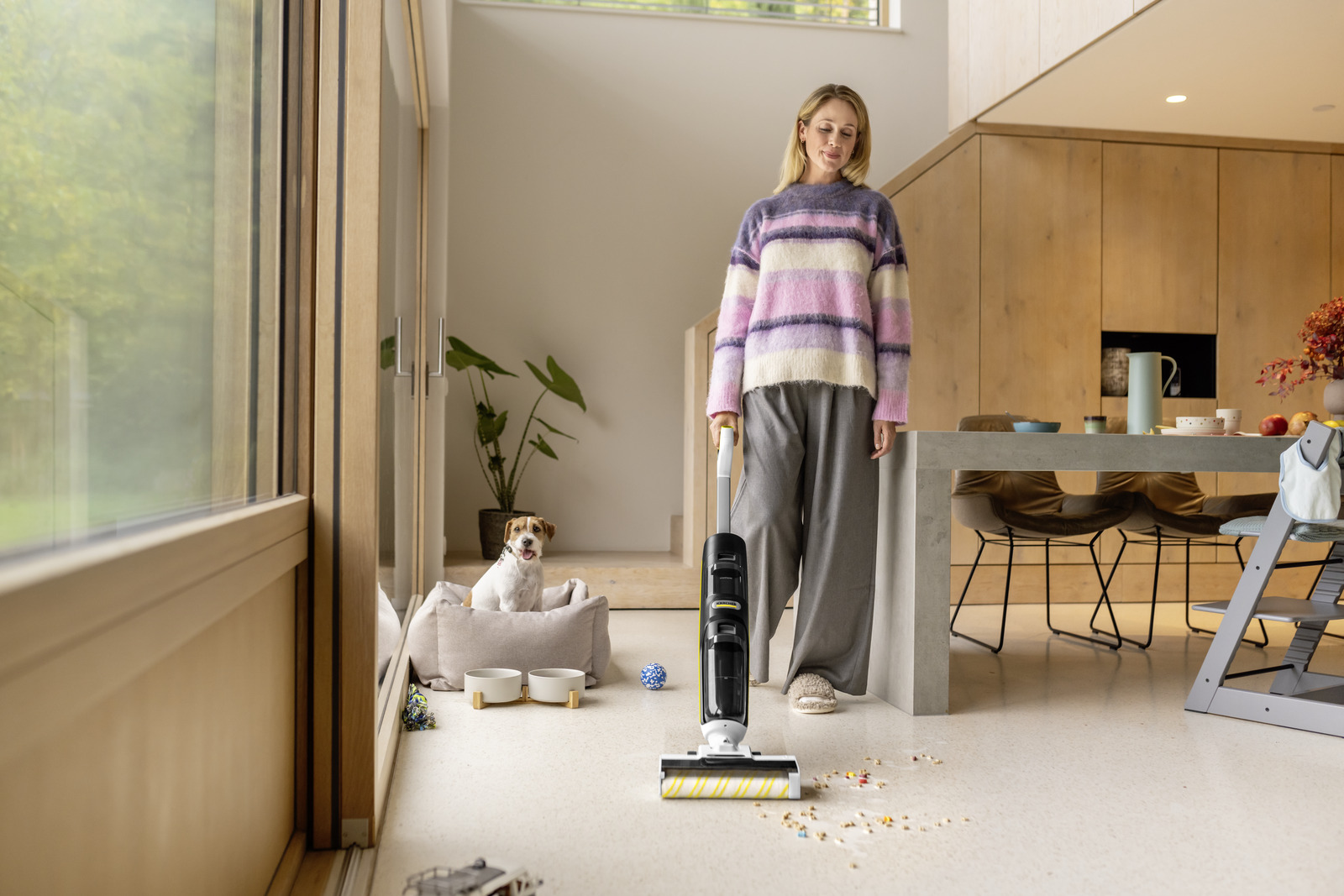 Een vrouw stofzuigt de vloer, met een hondenmand en een hond op de achtergrond. Een tafel en een keuken zijn ook zichtbaar.