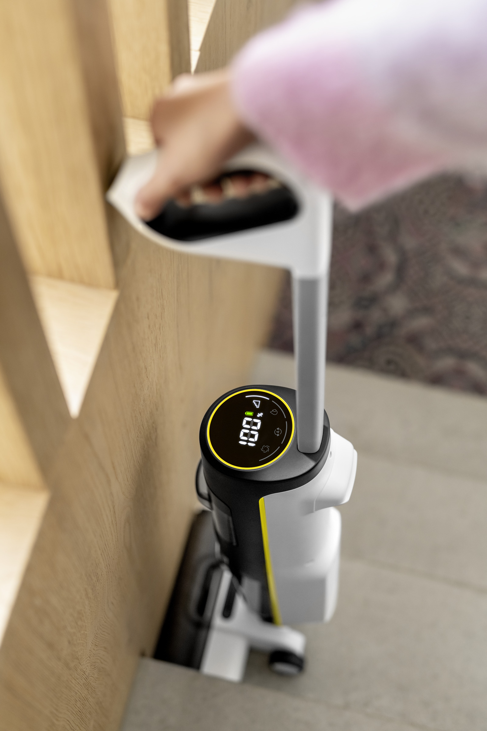 Een persoon houdt een wit en geel schoonmaakapparaat vast. Het apparaat heeft een digitaal display met 100%. Linkerzijde is een houten muur.