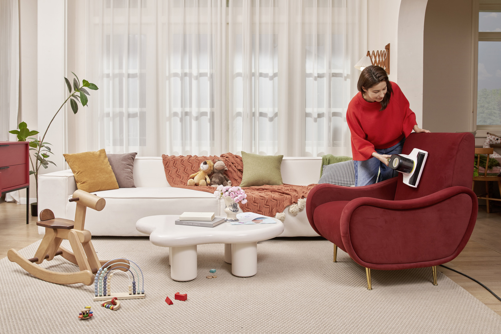 Frau in rotem Pullover saugt einen roten Sessel. Wohnzimmer mit weißem Sofa, Spielzeug, Couchtisch und Vorhängen.
