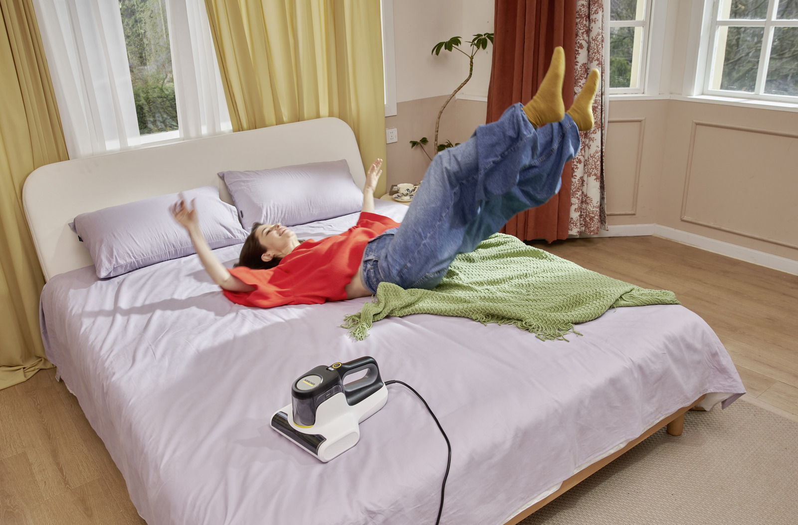 Frau in rotem Hemd und Jeans auf Bett mit Staubsauger. Gelbe Socken, grüne Decke. Vorhänge und Fenster im Hintergrund.