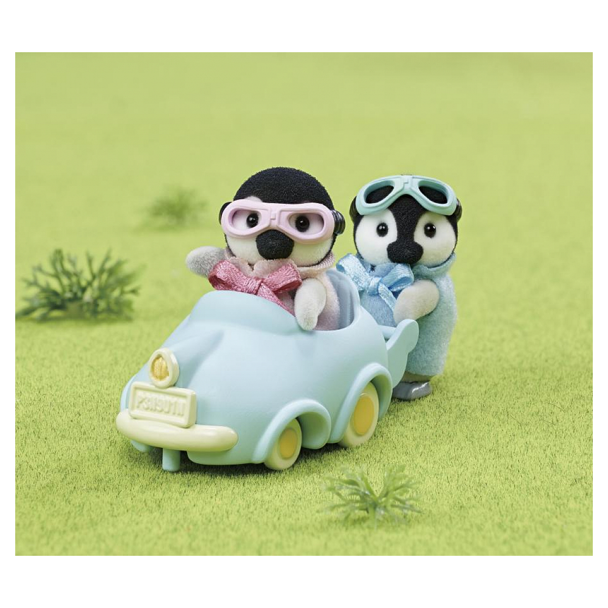 Dos juguetes de pingüino en un coche azul claro sobre hierba verde. Uno lleva gafas rosas, el otro una bufanda azul y gafas.