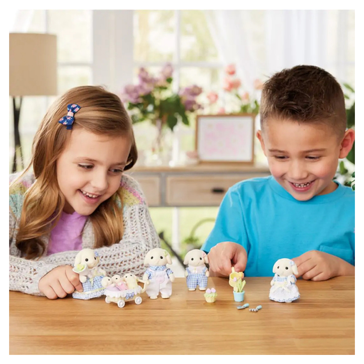 Dos niños sonrientes jugando con conejitos de juguete en una mesa de madera. El fondo incluye una ventana, flores y un mueble decorativo.