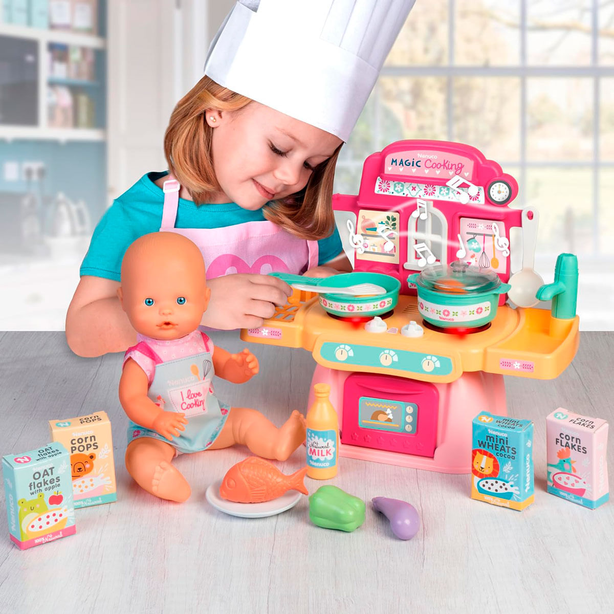 Una niña con un gorro de chef juega con una cocina de juguete y una muñeca. Hay juguetes de comida y cajas.