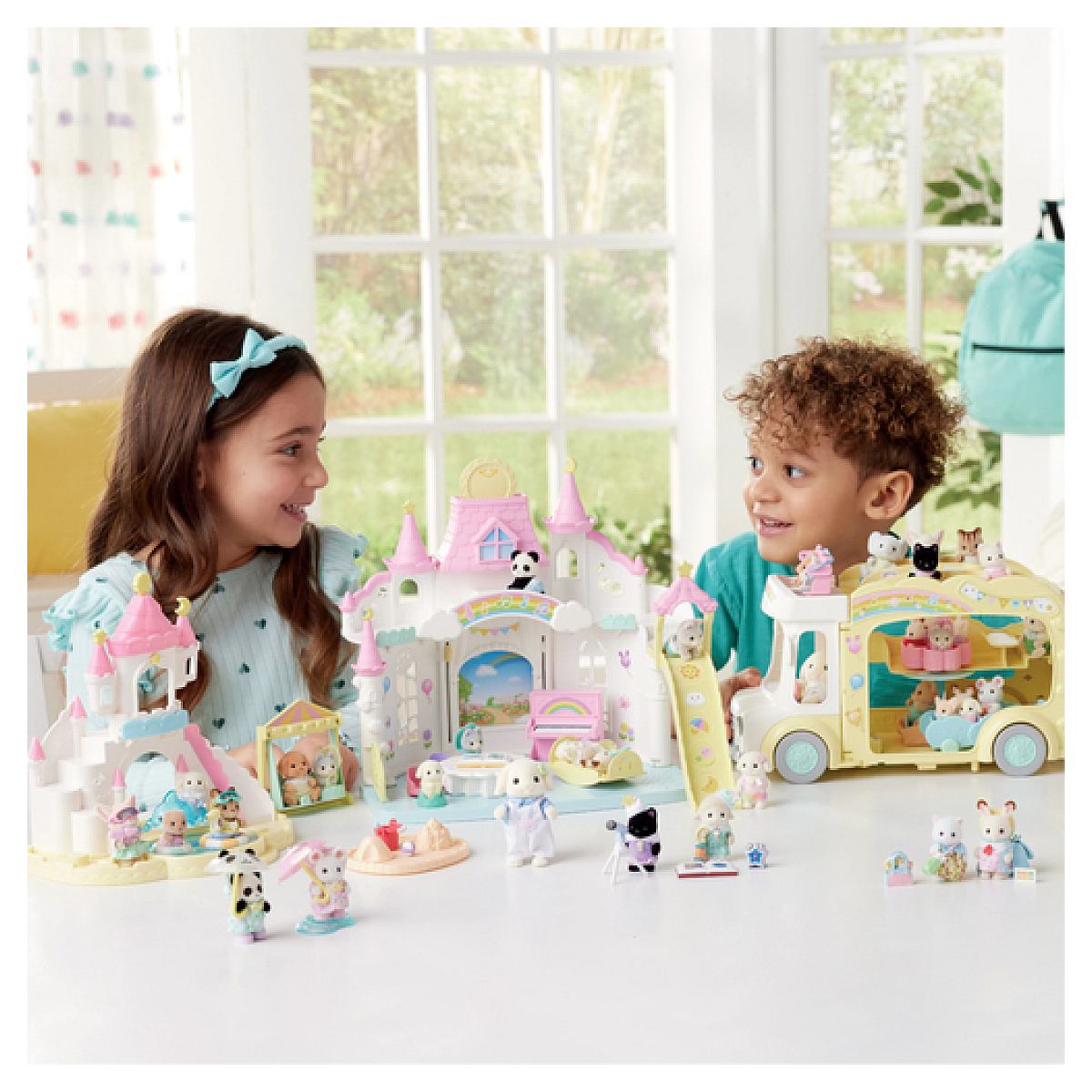 Dos niños sonríen a juegos de juguetes. Un castillo, un autobús y figuritas de animales están en una mesa blanca. El fondo es una ventana.