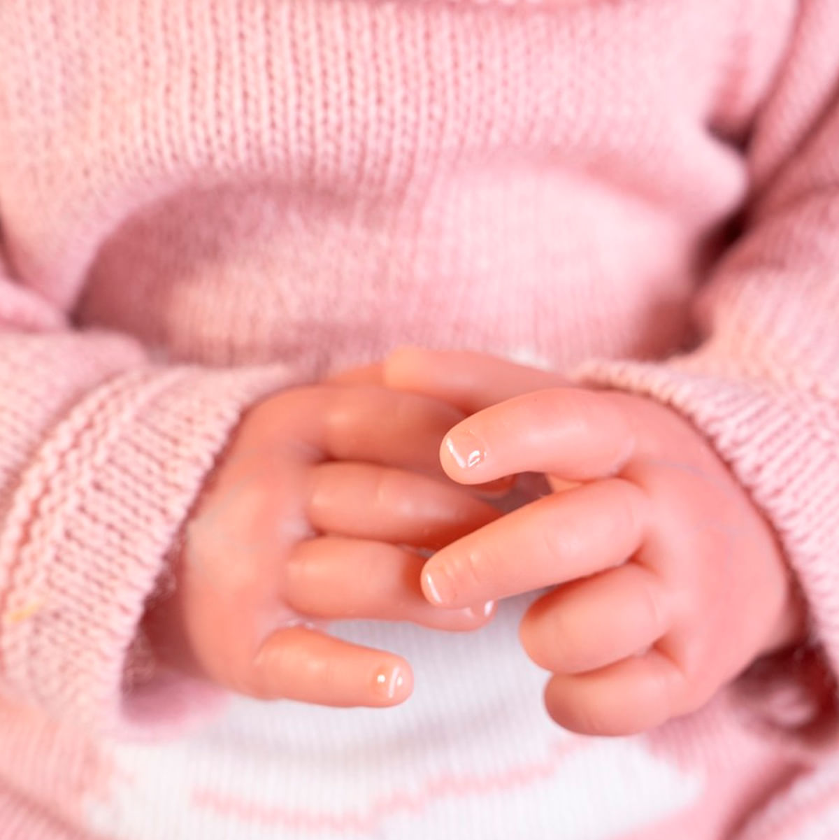 Primer plano de las manos de un bebé, juntas. El bebé lleva un suéter rosa. La piel es pálida, las uñas son visibles. El fondo es claro.