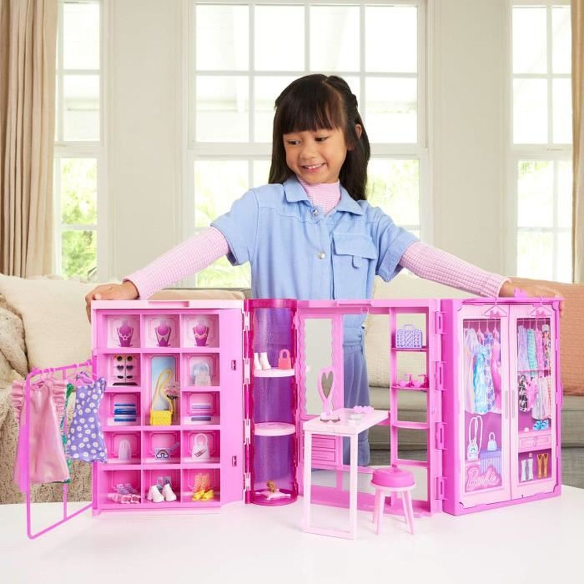 Una niña sonríe, abriendo un armario de Barbie rosa. El juguete tiene estantes, ropa colgante, un tocador y un taburete. Fondo blanco.