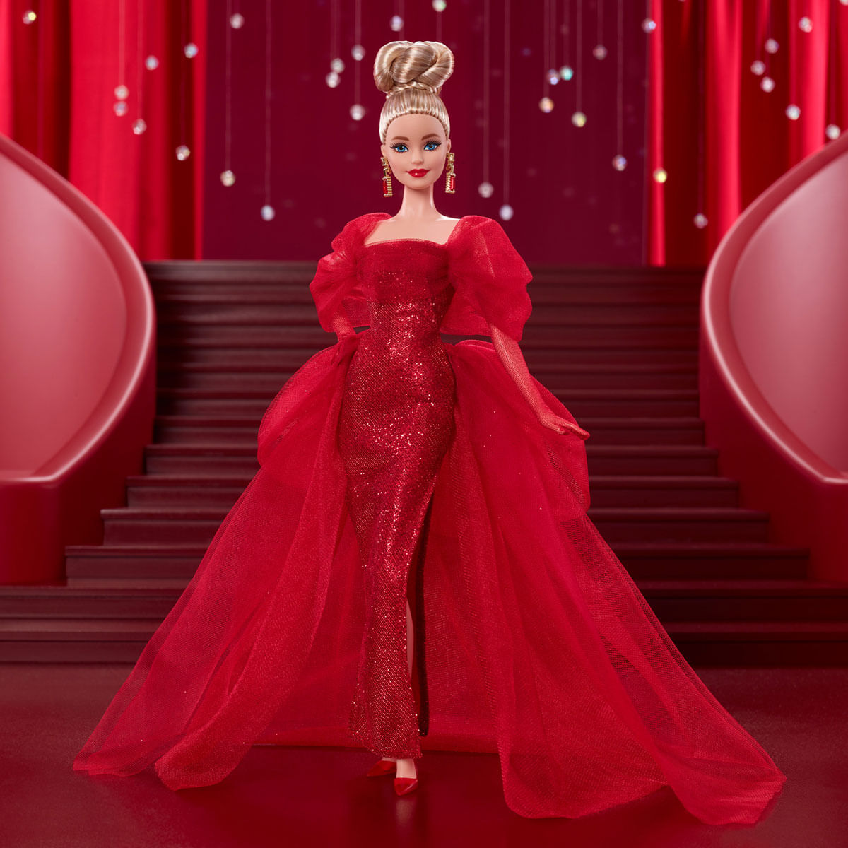 Una muñeca Barbie con un vestido rojo con cola, de pie en un escenario rojo con escaleras en el fondo y luces brillantes.