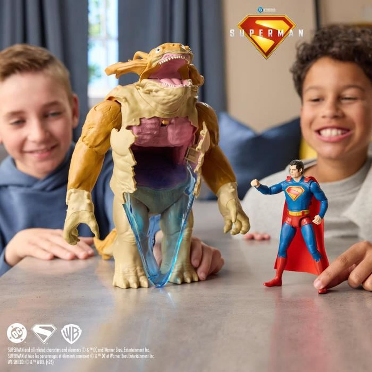 Dos niños sonriendo a un monstruo de juguete y una figura de Superman. El monstruo tiene el pecho abierto. Superman está delante de él.