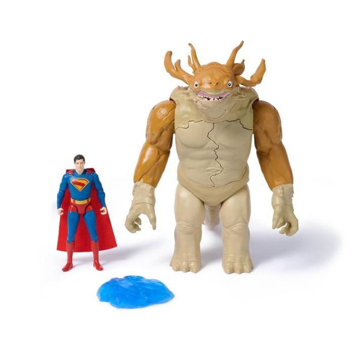La figura de Superman está junto a un gran monstruo de juguete con cuernos y una mancha azul. El fondo es blanco.