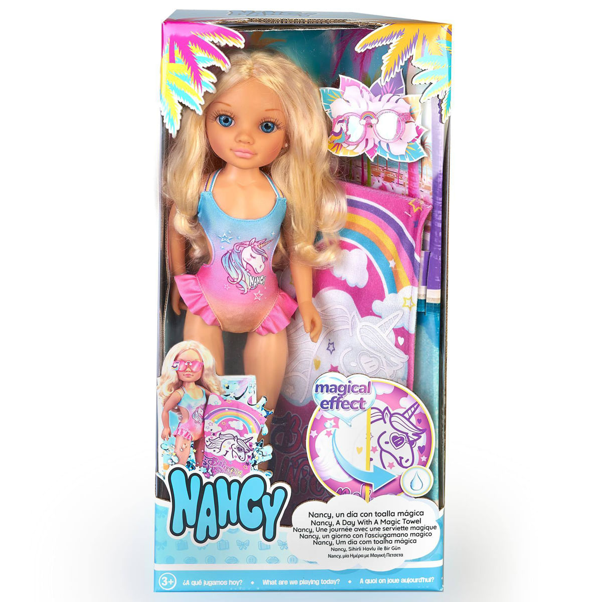 Una muñeca Nancy en una caja. Lleva un bañador con un unicornio. Hay una toalla con un arcoíris y un unicornio.