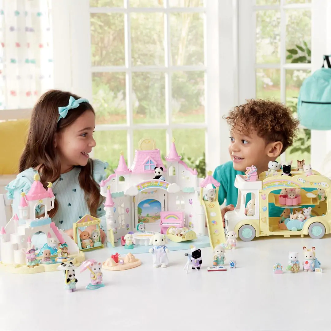 Zwei Kinder lächeln an einem Tisch mit Spielzeugschlössern und einem Bus. Kleine Tierfiguren spielen um das Spielzeug herum. Im Hintergrund ist ein sonniges Fenster.