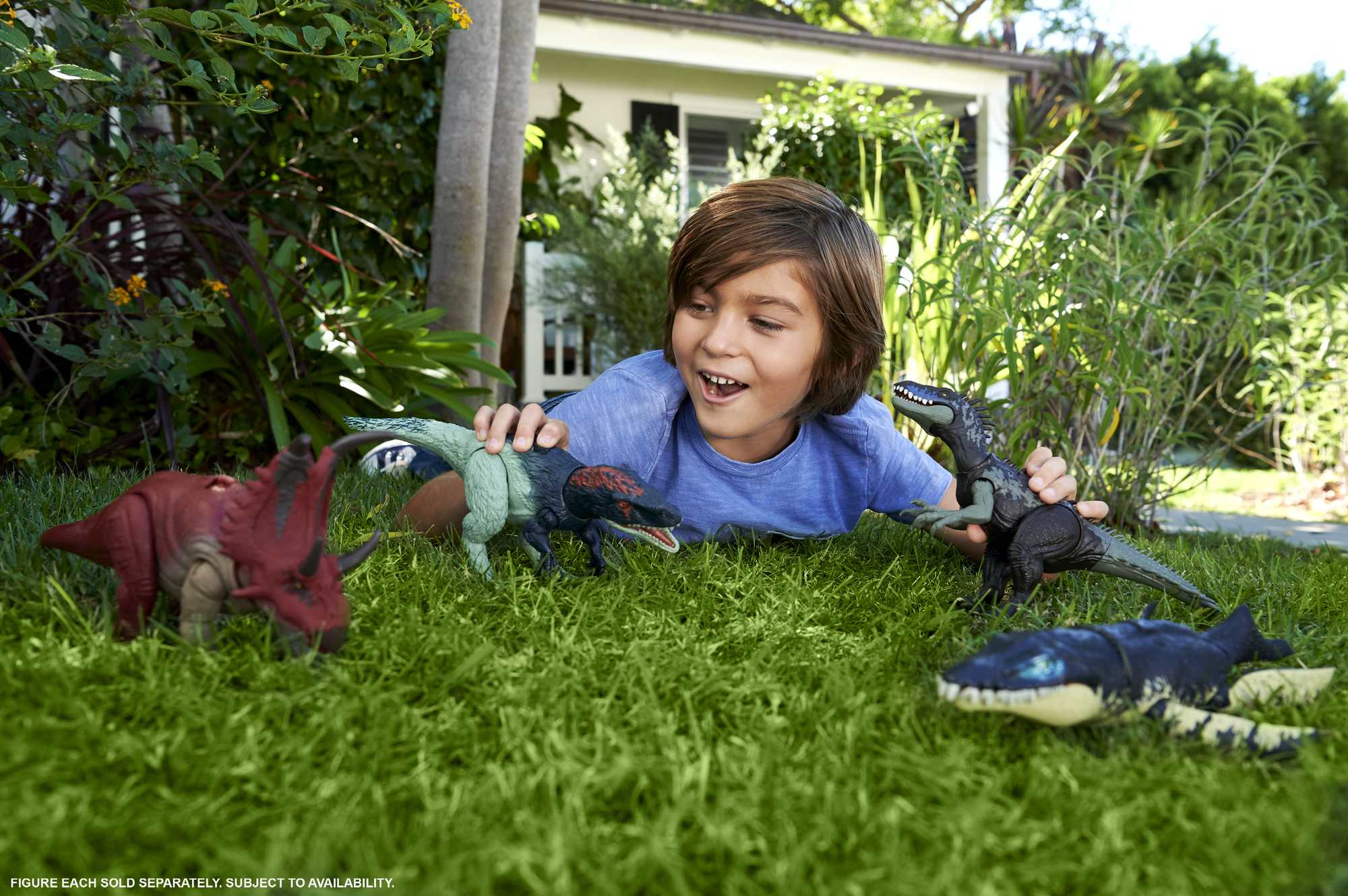 Un niño sonríe, rodeado de juguetes de dinosaurios sobre hierba. Una casa y plantas al fondo.