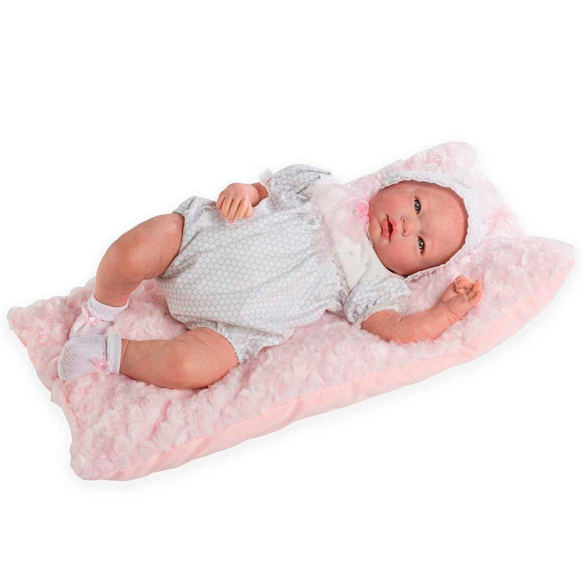 Una muñeca bebé yace sobre una almohada rosa. Lleva un pelele blanco, gorro y calcetines. La muñeca tiene ojos azules y mira hacia arriba.