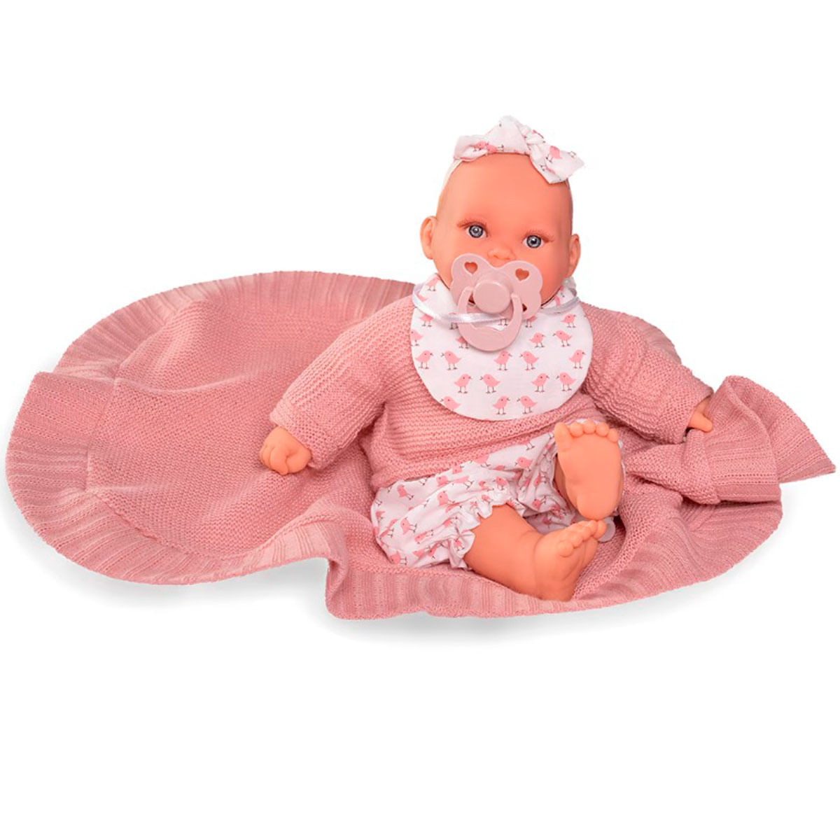 Una muñeca bebé, con suéter rosa, pantalones cortos y babero con estampado de pájaros, está sobre una manta rosa. Tiene un chupete y un lazo.