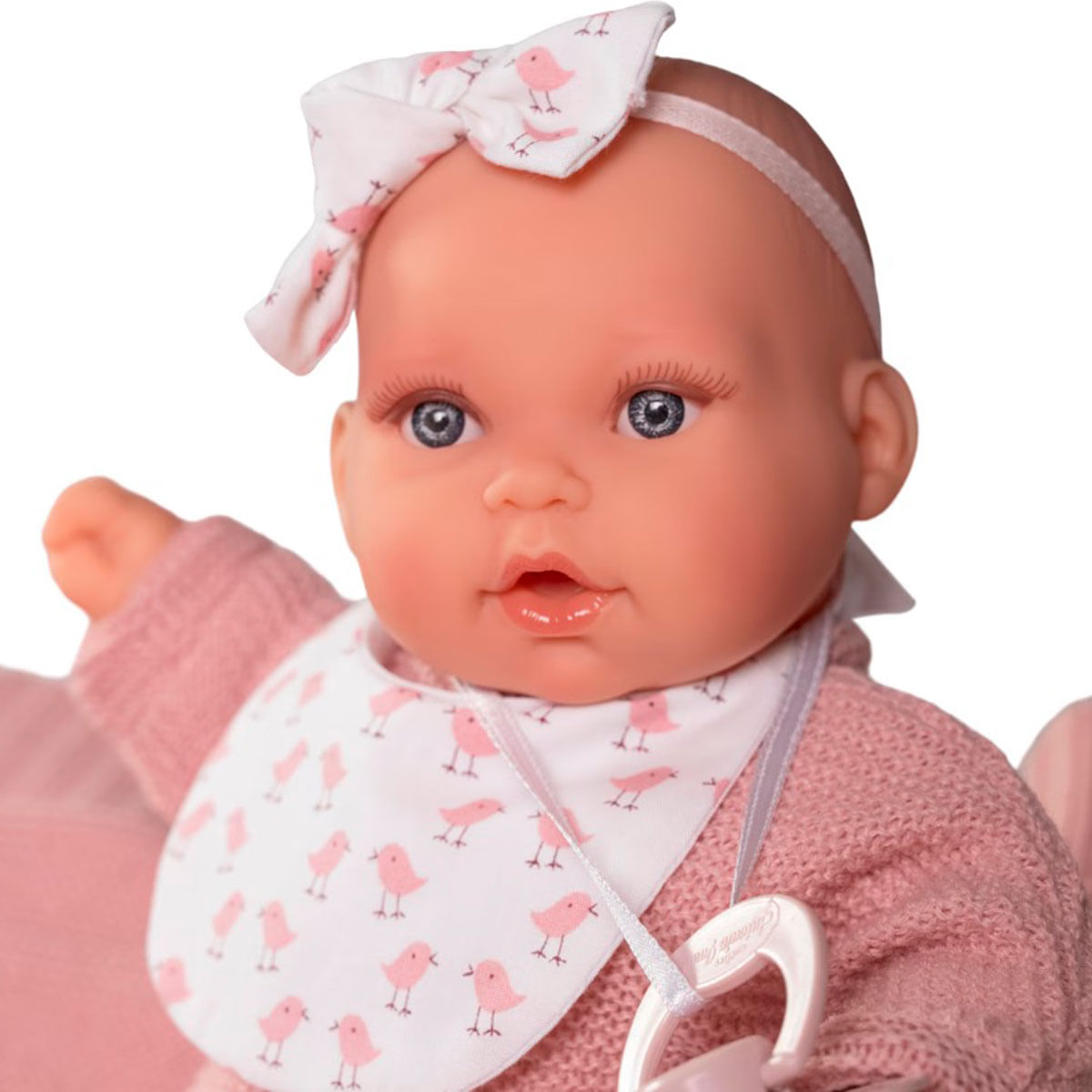 Una muñeca con suéter, babero y lazo rosas. El babero y el lazo tienen dibujos de pájaros. La muñeca tiene ojos azules.