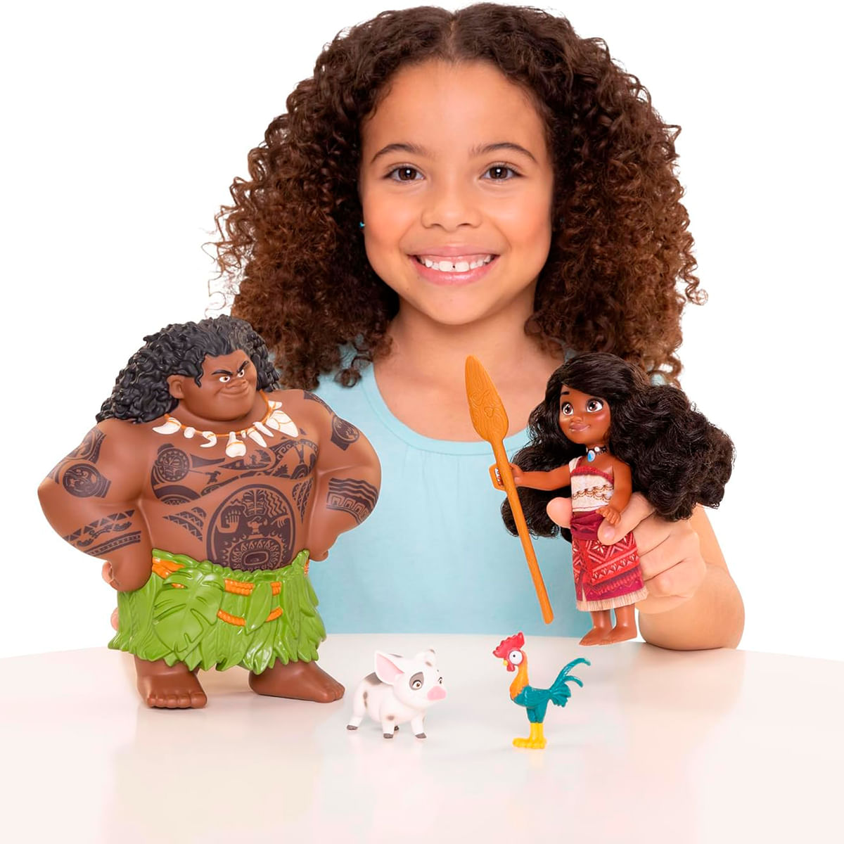 Una niña sonríe, sosteniendo una muñeca Moana y muñecos de Maui, un cerdo y un gallo. Los muñecos están sobre una superficie blanca.