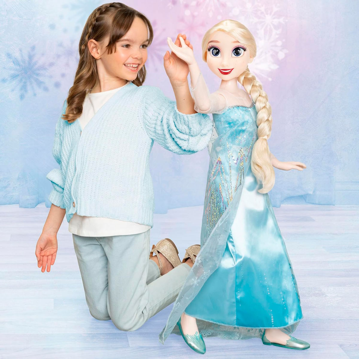 Una niña se arrodilla, tocando la mano de una muñeca Elsa grande. Ambas visten de azul. Fondo con copos de nieve.