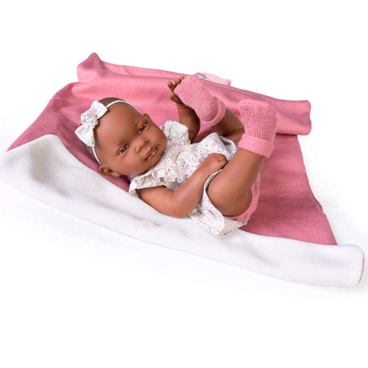 Una muñeca bebé con piel oscura está acostada sobre una manta rosa. Lleva un vestido floral y calcetines rosas. Tiene una diadema blanca con un lazo en la cabeza.