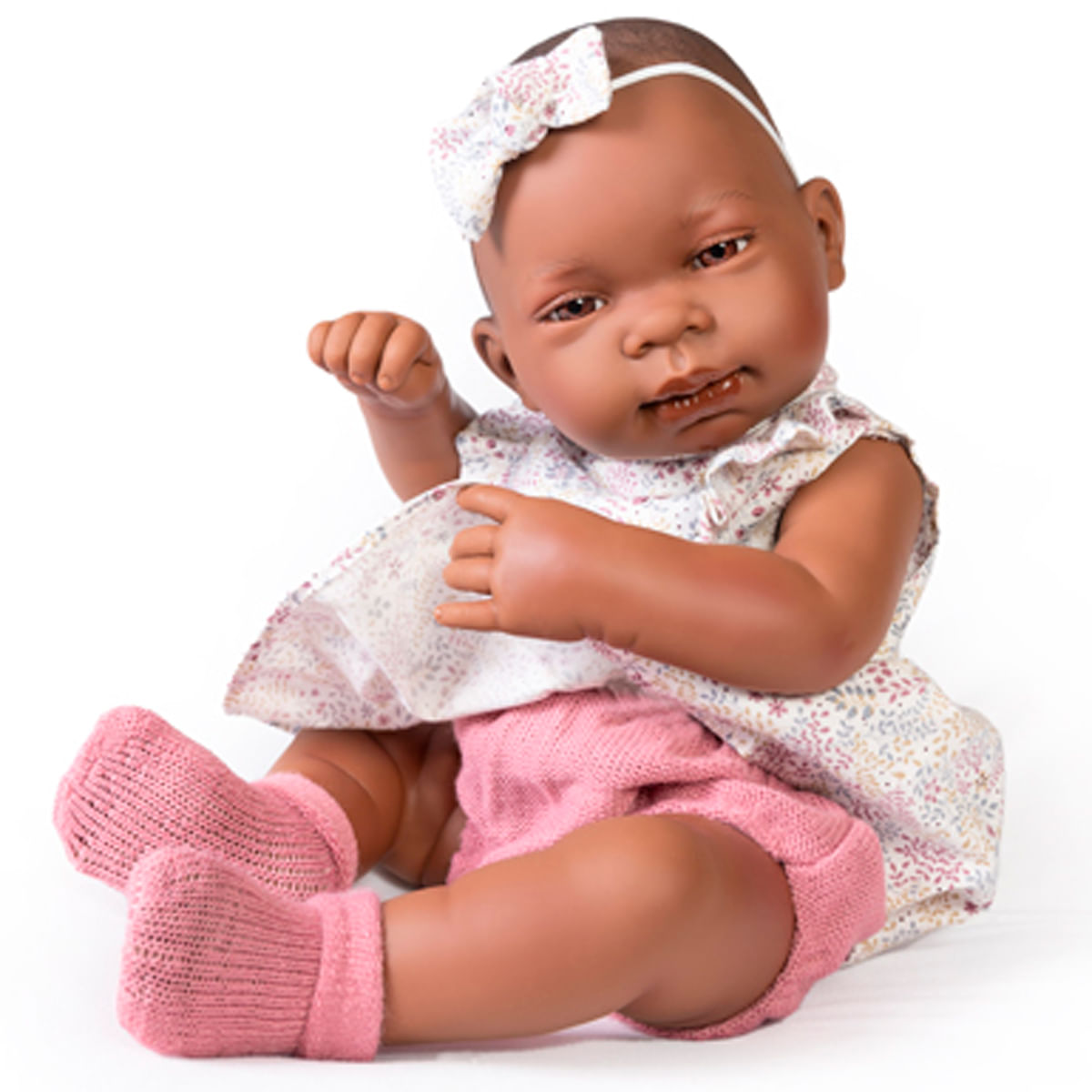 Una muñeca bebé de piel oscura, con vestido floral, pantalones cortos rosas, calcetines y un lazo. Sentada sobre fondo blanco.