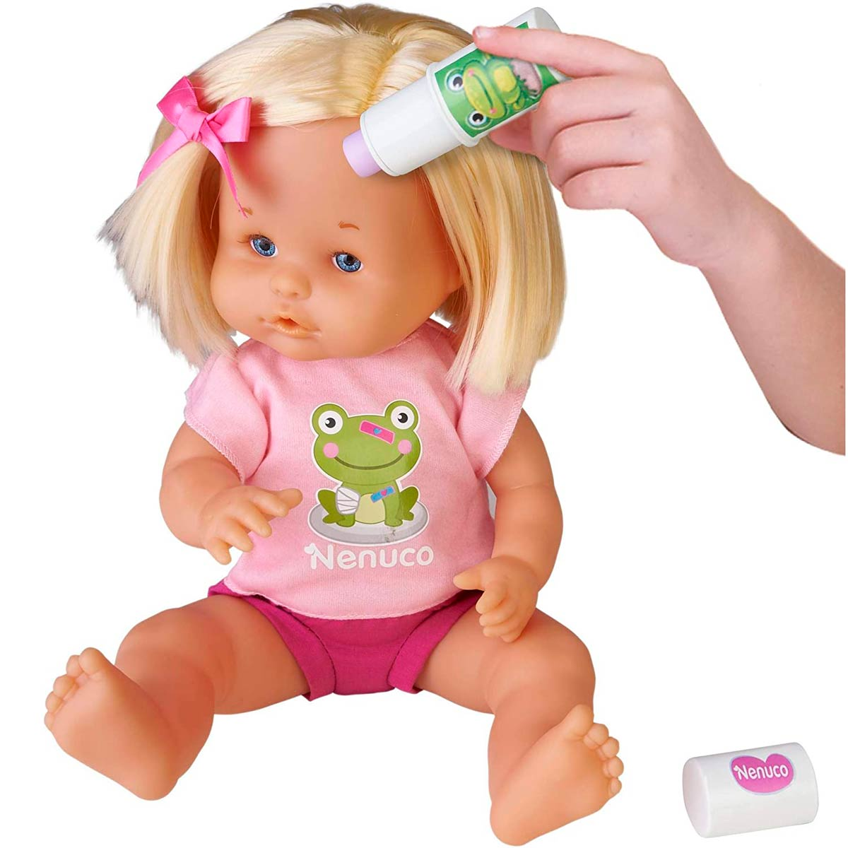Una muñeca rubia con una camisa rosa recibe crema aplicada por una mano. Una rana está en la camisa. Hay un tubo de crema.