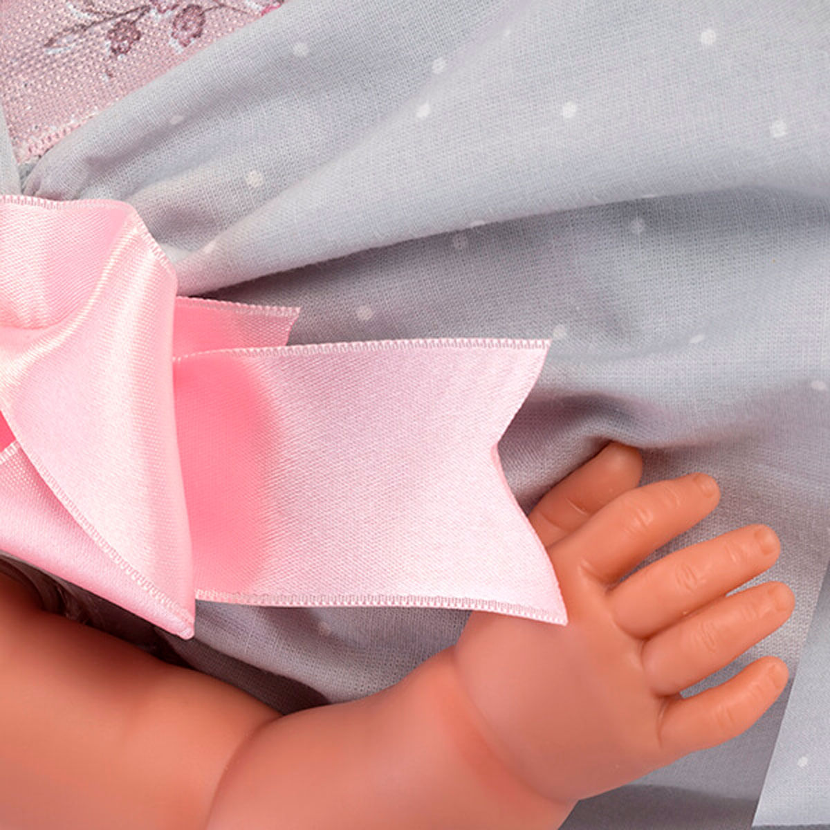 Primer plano de una mano de muñeca, un lazo rosa y tela gris con puntos. La piel de la muñeca es pálida. El lazo es prominente.