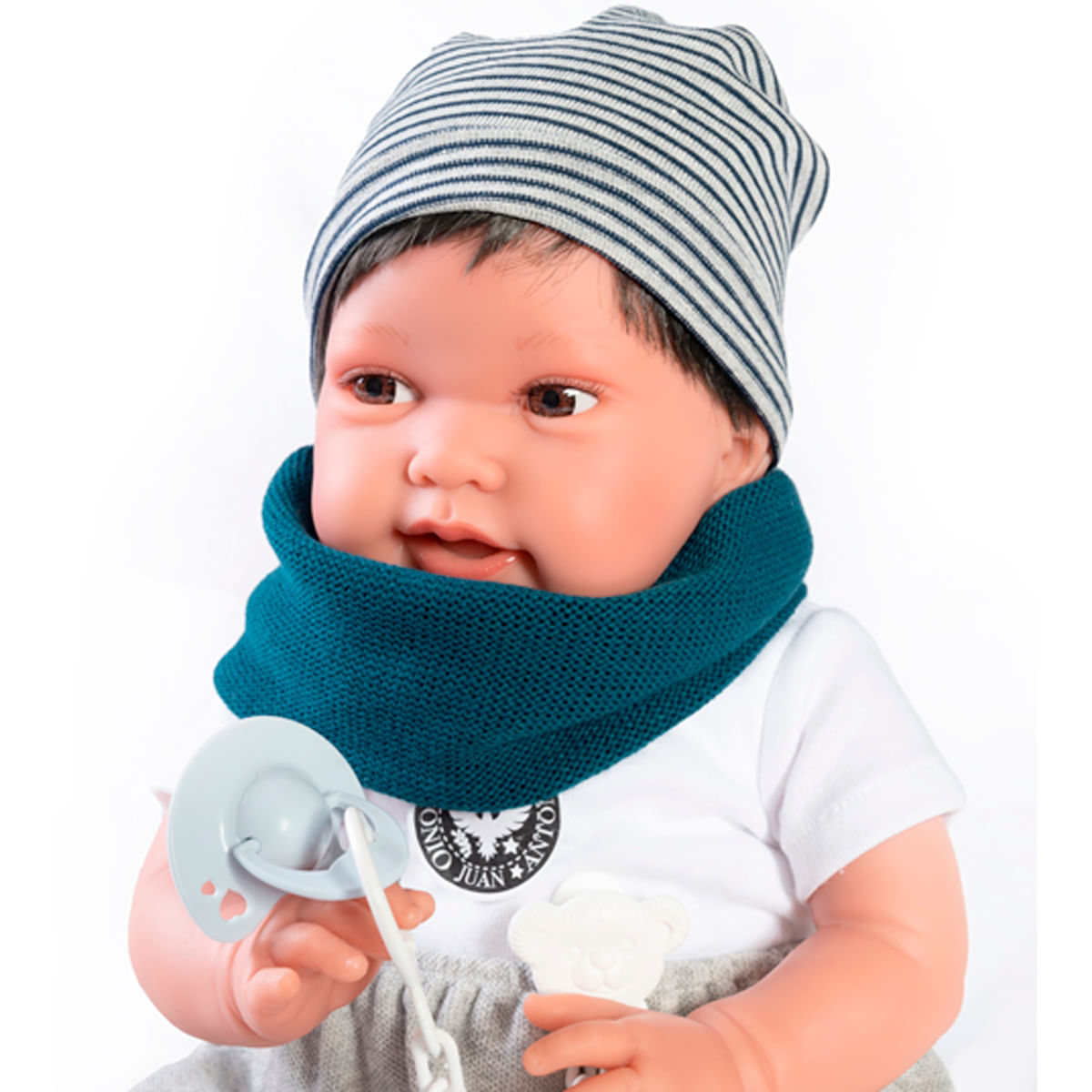 Una muñeca con gorro a rayas, bufanda turquesa y camisa blanca, sosteniendo un chupete. Tiene pelo oscuro y ojos marrones. El fondo es blanco.