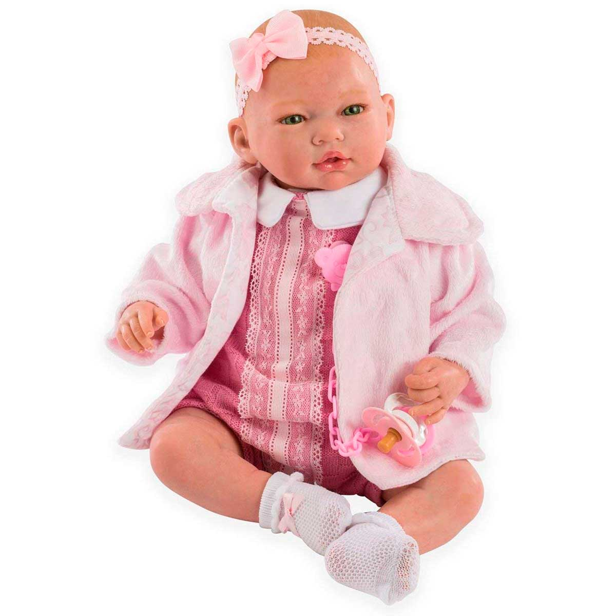 Una muñeca bebé con ropa rosa, diadema y calcetines blancos, sosteniendo un chupete. La muñeca está sentada, sobre fondo blanco.