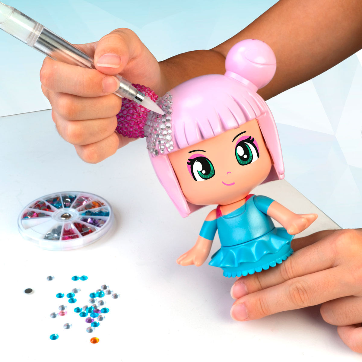 Una mano decora el pelo de una muñeca con pedrería usando una herramienta. Un contenedor de gemas y pedrería suelta está cerca.