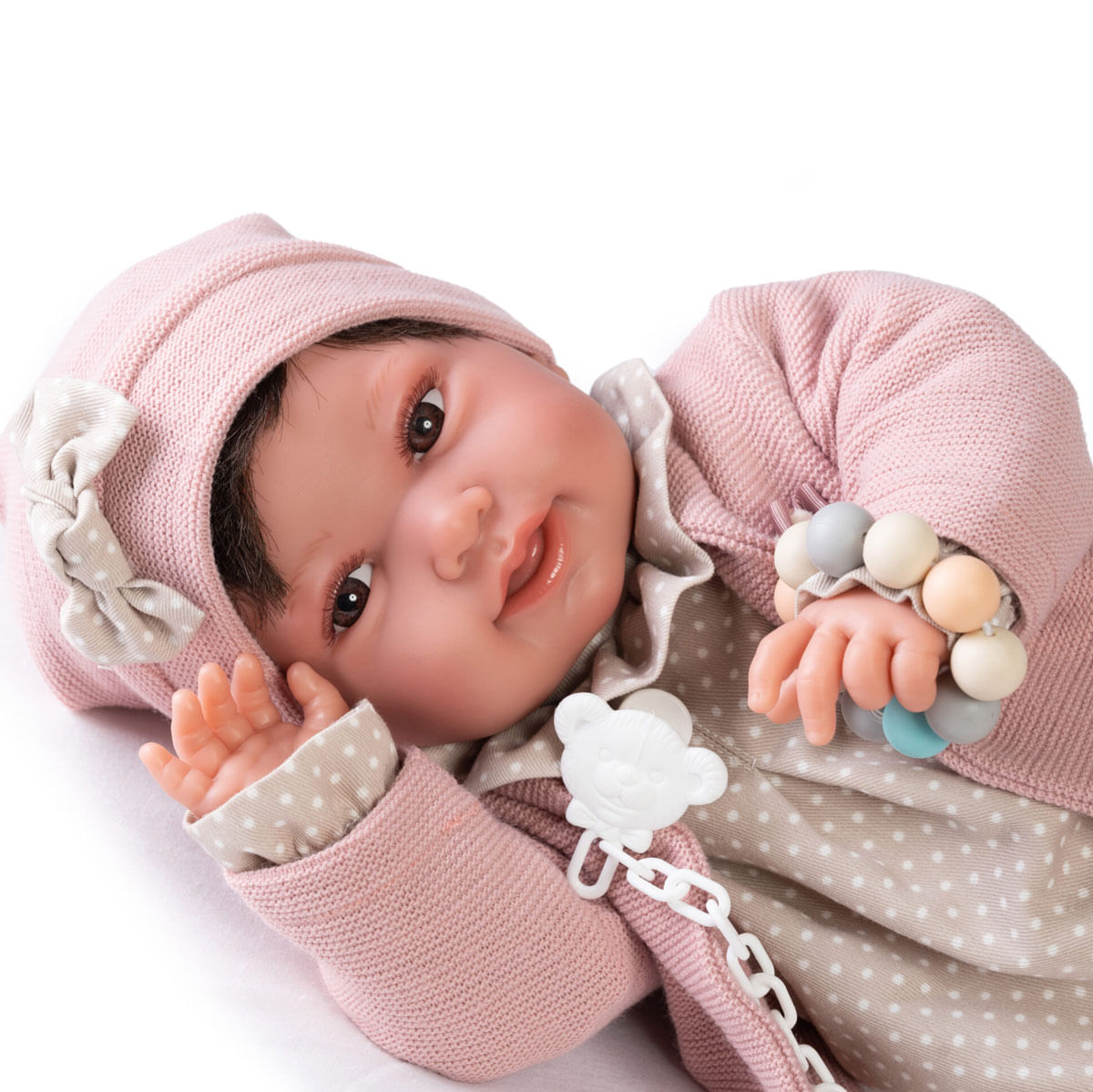 Una muñeca bebé con ropa y gorro rosa yace sobre blanco, sosteniendo una pulsera de cuentas. Tiene ojos marrones y un chupete.