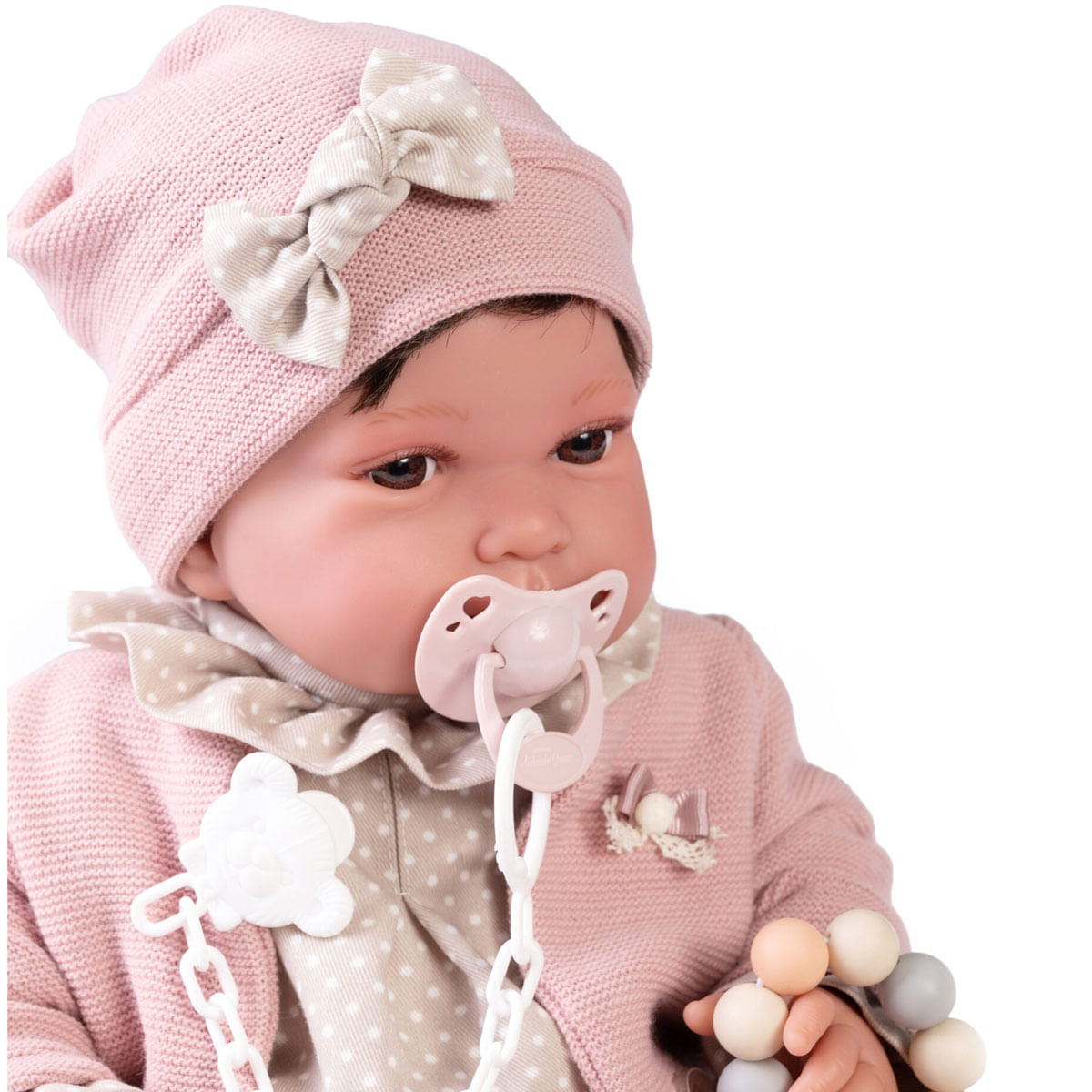 Una muñeca con gorro rosa y lazo, suéter rosa y chupete. La muñeca sostiene un juguete de cuentas.