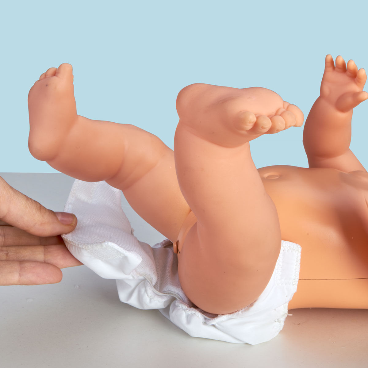 Se ven las piernas y el cuerpo de una muñeca. Una mano ajusta el pañal. La muñeca está sobre una superficie blanca con fondo azul.